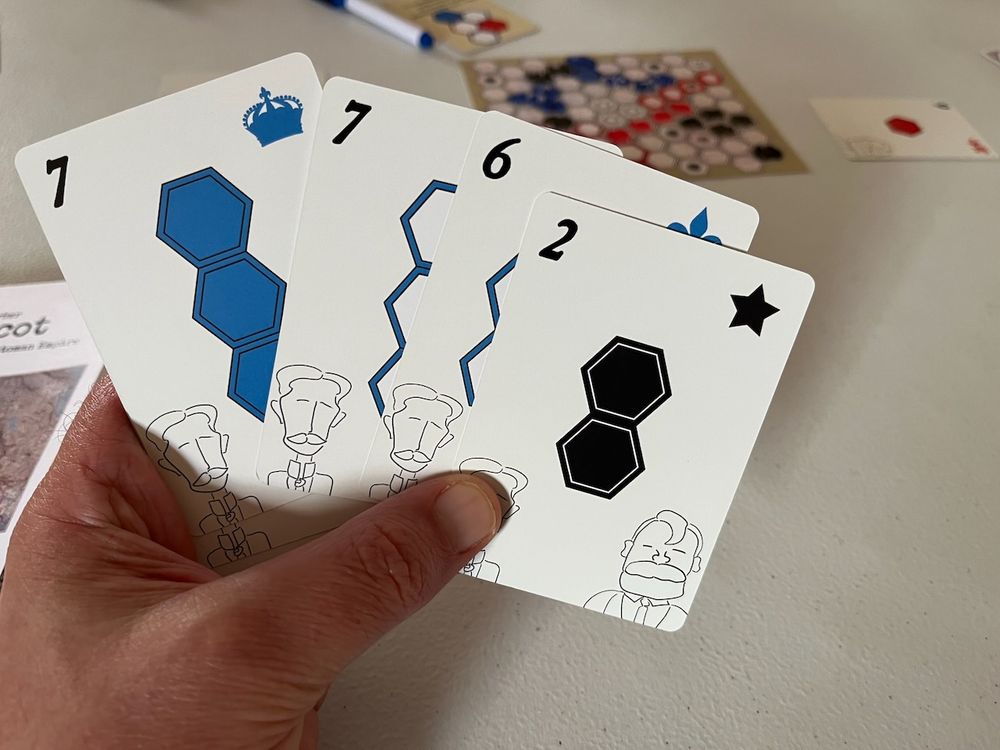 Shadowed hand holding up four cards from Sykes-Picot: The Secret Treaty to Partition the Ottoman Empire. The cards have numerals at upper left, symbols at upper right (black star, fleur, blue crown) and hex configurations in the middle. Line illustrations of 19th Century style European men are at the bottom of the cards.