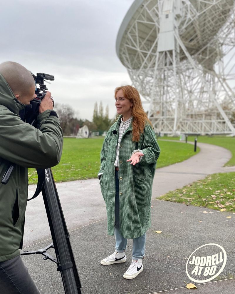A photo of musician Hannah Peel, a woman with red hair and a green coat, being filmed in front of the Lovell Telescope.