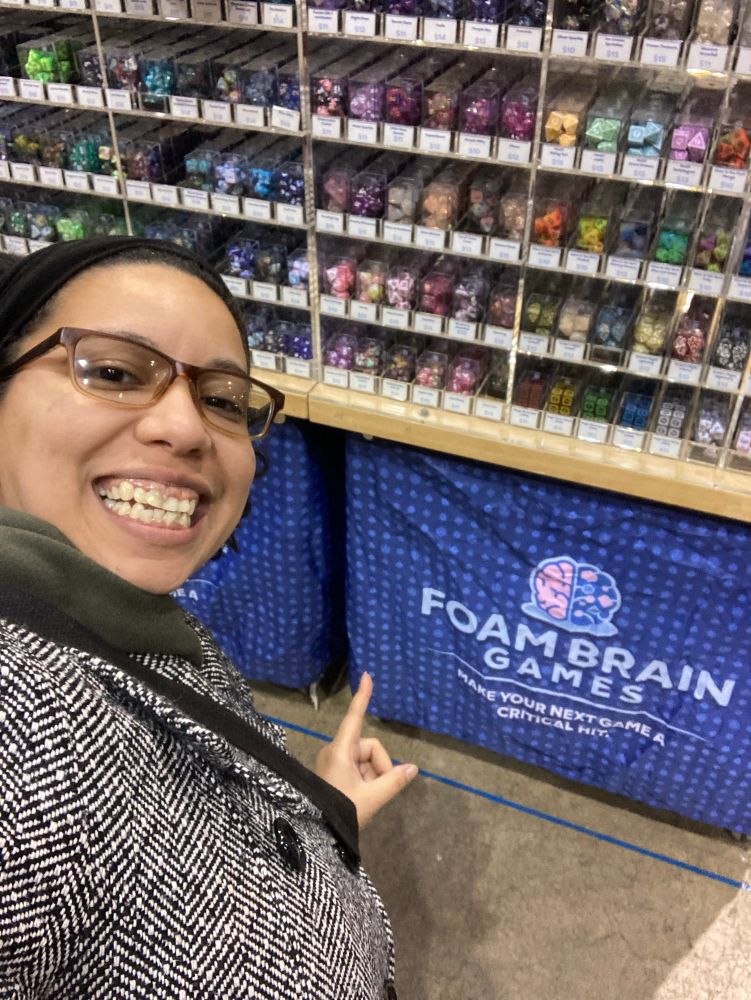 Raquel posing in front of a display case full of various RPG dice sets, pointing at the Foam Brain Games logo 