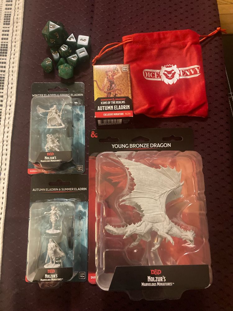Various items laid out on a table: miniatures for Eladrins of all 4 seasons, mini for a Young Bronze Dragon, a dice bag with Dice Envy written on it, and a 10 piece set of green & gold gaming dice 