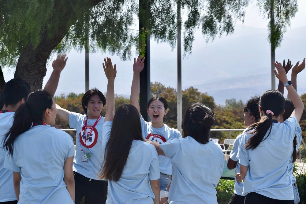 Group of Red Cross youth in matching t-shirts standing in a circle, some with arms raised, in a park with trees in the background. 