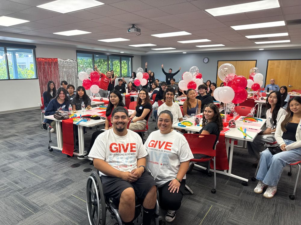 Group of students at tables with red and white balloons. Two individuals are seated in front of the students. 