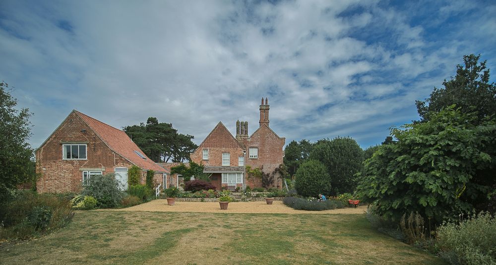 A charming English country house with distinctive red brick construction and terracotta tile roofing, featuring multiple chimneys and traditional architecture. The property sits at the edge of a well-maintained but slightly sun-dried lawn, surrounded by mature trees and lush hedgerow borders. The main house shows Arts and Crafts-style influences with its steep gabled roofs and prominent brickwork, while an attached wing extends to the left. The garden appears established and peaceful, with garden furniture visible in the distance, all set against a partly cloudy blue summer sky.