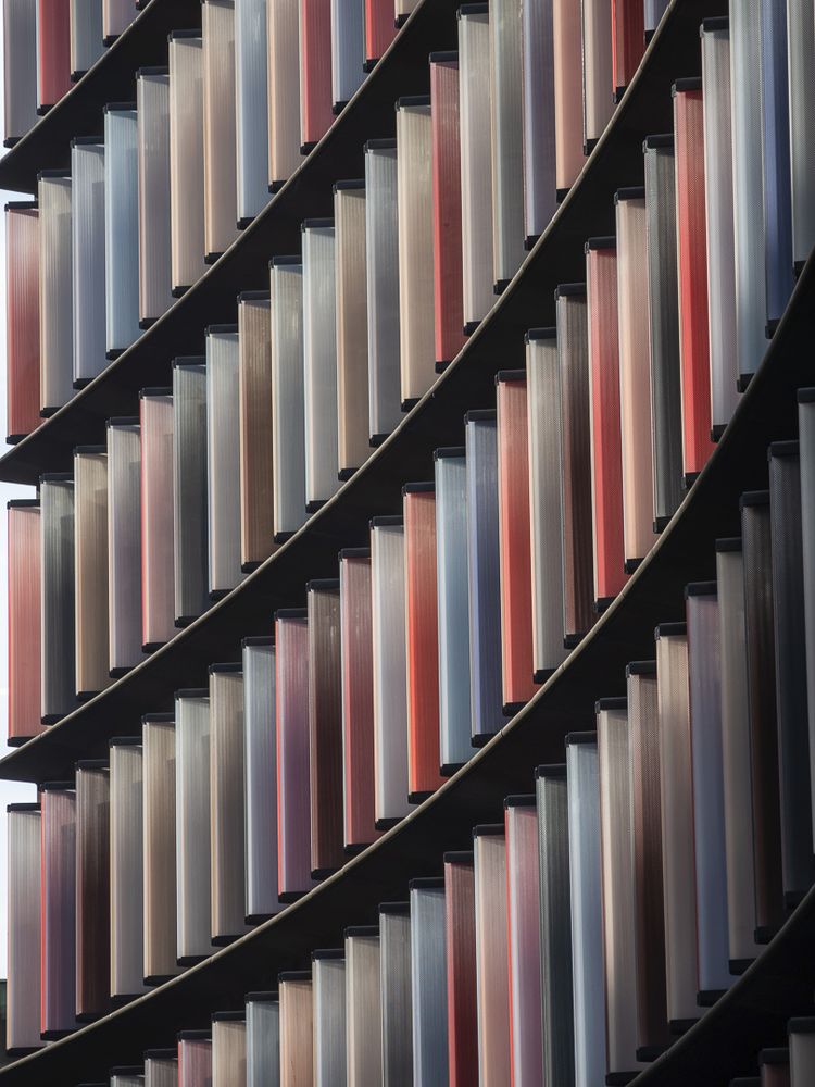 Architectural detail showing diagonal rows of vertical metal louvers or fins in varying muted tones of grey, beige, coral, and rust, creating a rhythmic geometric pattern across a building facade. The elements are arranged in repeating stepped formations that run from upper left to lower right, with strong shadows emphasizing their dimensional depth and industrial aesthetic.