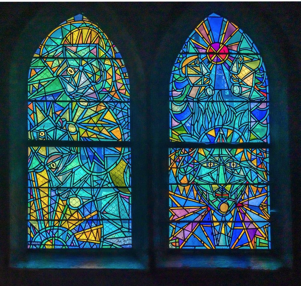 Two tall arched stained glass windows featuring abstract geometric designs in vibrant blues, teals, yellows, oranges, and purples. The left window contains flowing angular patterns with eye motifs scattered throughout. The right window displays a radial sunburst pattern at the top with a central cross motif, and geometric eye symbols in the lower sections. Both windows are divided into three horizontal sections by dark metal frames and are set against a dark wall, creating a luminous effect