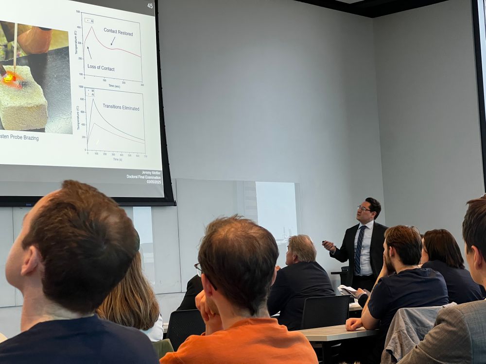 Picture of Jeremy Mettler presenting his PhD defense in a packed room of people (it was so full we had people sitting on the floor and standing around the edges). 