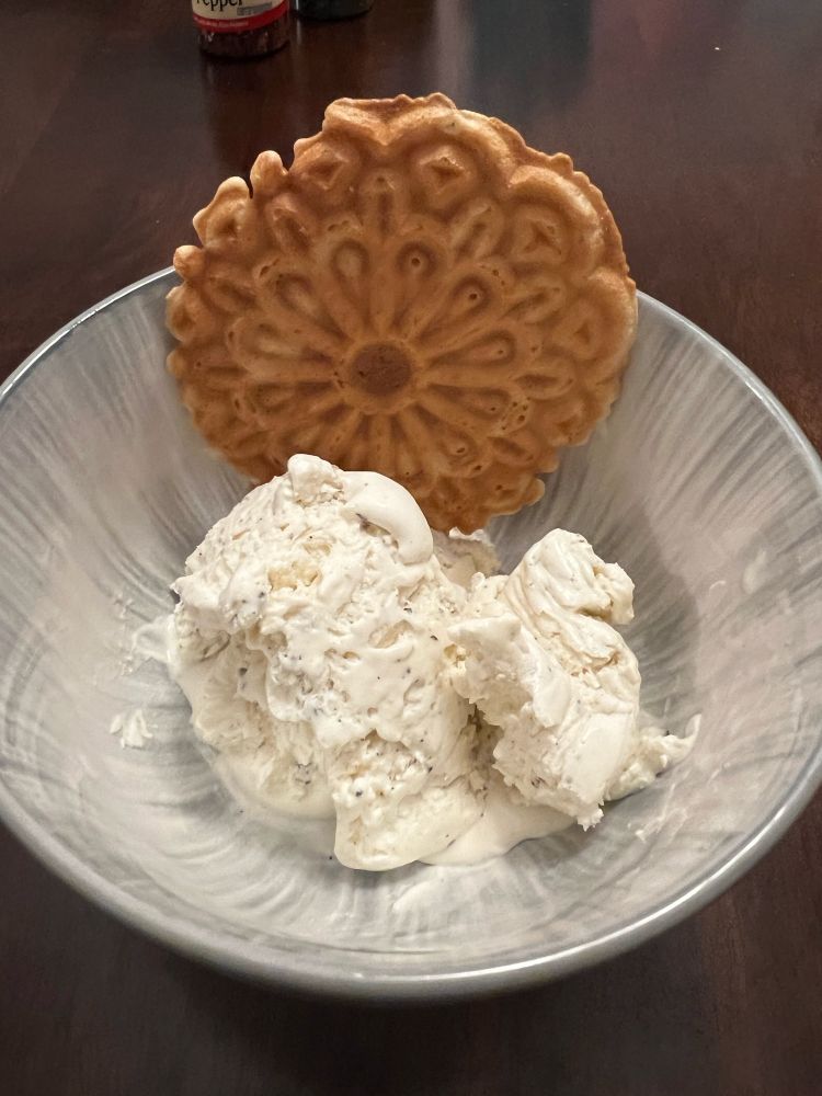 A bowl with a single scoop of ice cream that has flecks of black pepper visible throughout. Nestled between the ice cream and the bowl is a thinly-pressed, lemon waffle cookie with rough edges.