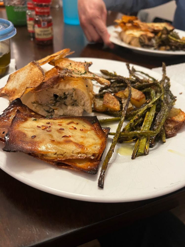 Dinner plate with thin strips of fried potato atop a spinach and cheese stuffed chicken breast. In front of the chicken is a stack of potato sheets stuffed with cheese and spinach with red pepper flakes on top. Filling the rest of the plate is roasted asparagus with some roasted potatoes (we used the leftover bits from the potato sheets to make little quarters).