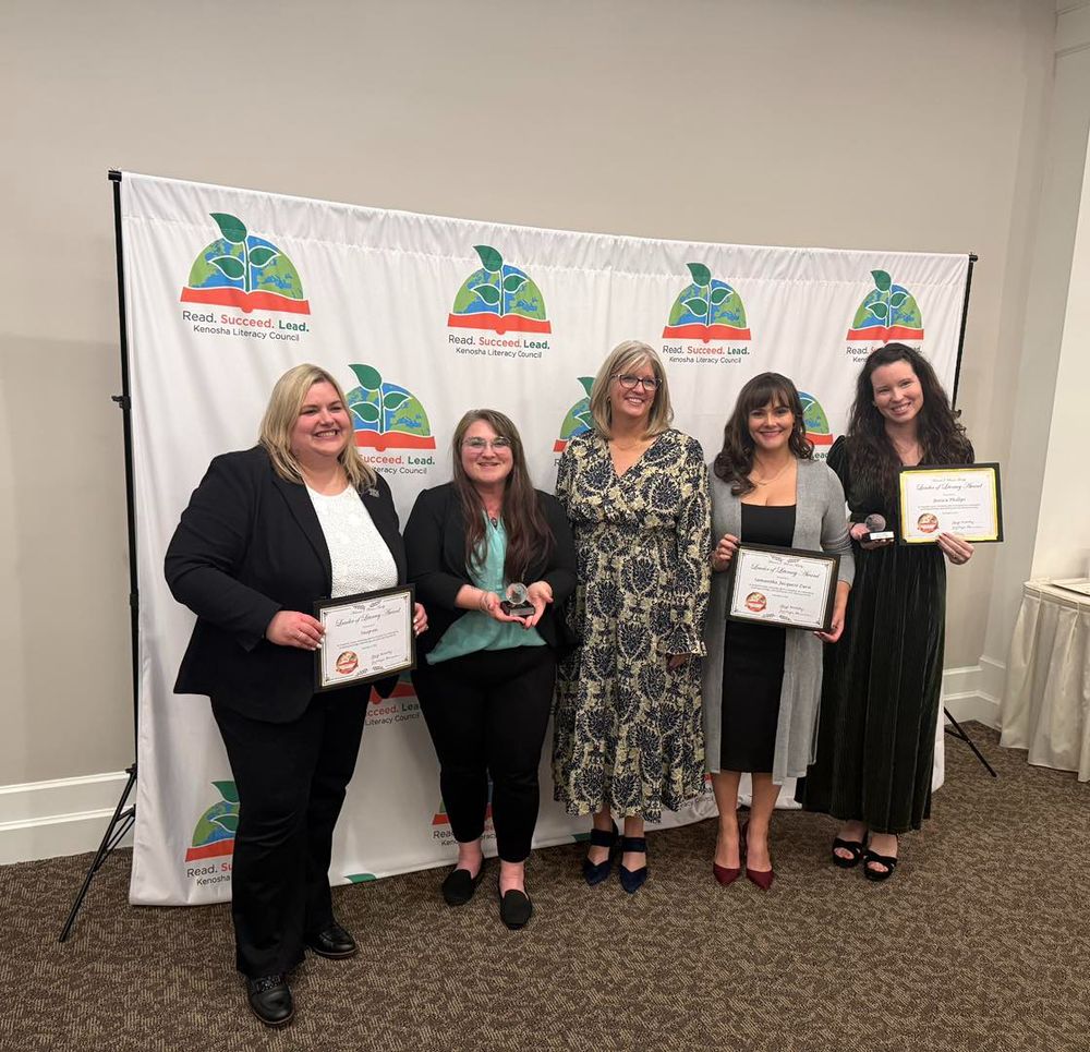 A photo of the KLC's winners during their event, including Sam, the owner of Blue House Books, in the long grey sweater over a black dress, holding up their awards. 