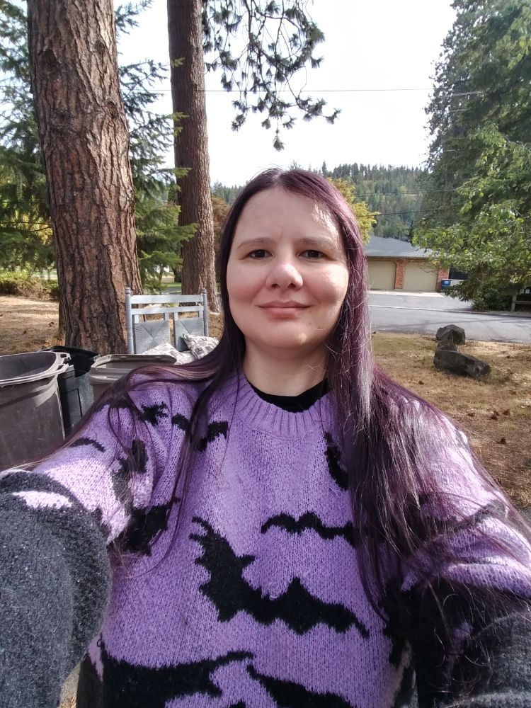 Author, Brooklyn Ann, (a short, middle-aged white woman with long purple hair) wearing a purple sweater with black bats on it. There are pine trees and mountains in the background. 