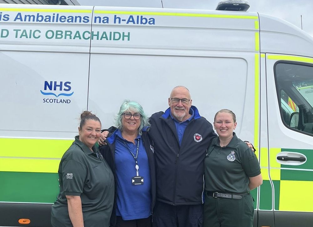Sam Grieve, Mary and John Kinninmonth, and Laura Cameron standing together, smiling, in front of a parked ambulance vehicle.