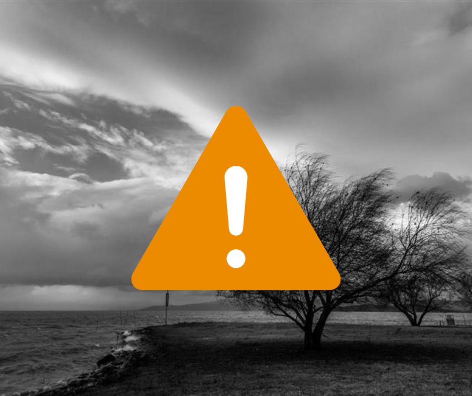 Dark storm clouds gather over a windy lakeside with bare trees bending in the strong breeze. An amber warning symbol is overlaid the top.