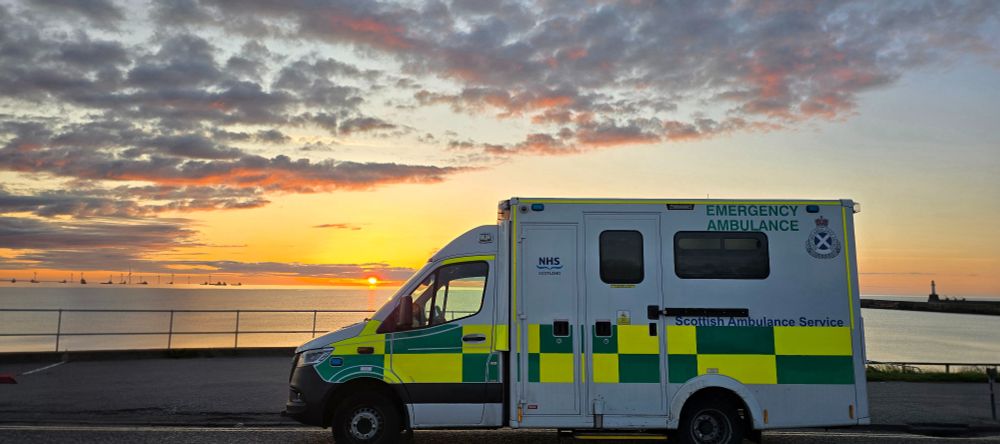 An ambulance parked by the coastline of a beach with a sun rising out of the sea in the distance.