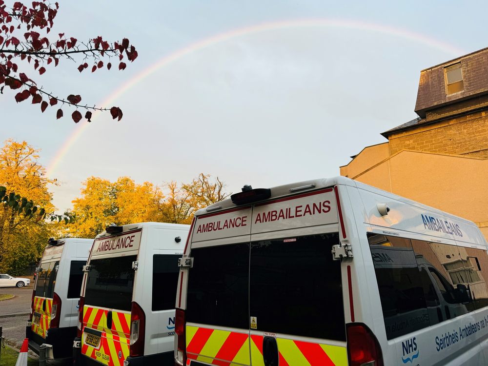 A vibrant rainbow arches over two parked ambulances near autumn trees and a beige building under a cloudy sky.