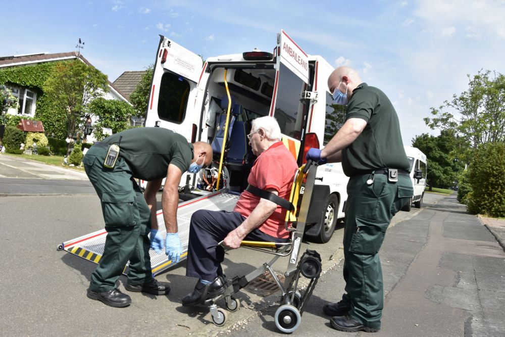 Two ambulance care assistants helping an elderly patient in a wheelchair into the back of an patient transport ambulance vehicle.