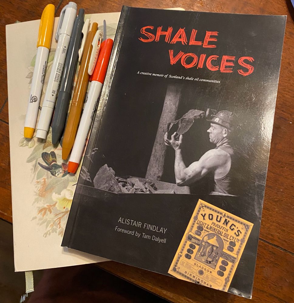 The book Shale Voices by Alistair Findlay lies on a wooden table with an array of pens in red, golden-yellow, and grey.
