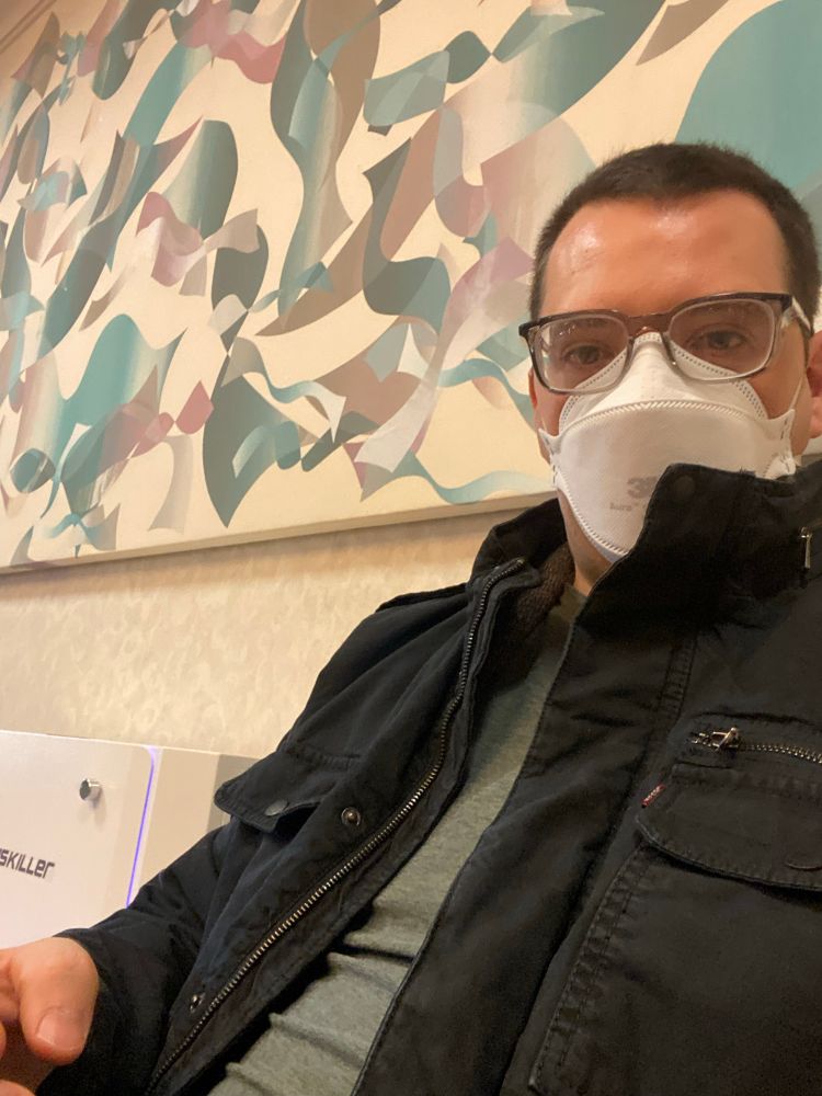 Photo of me, a white man with brown hair and glasses, sitting in a chair against a wall with a generic abstract painting. I’m wearing stoggles and a 3M Aura N95. Clothes, too—a black jacket over a green shirt 