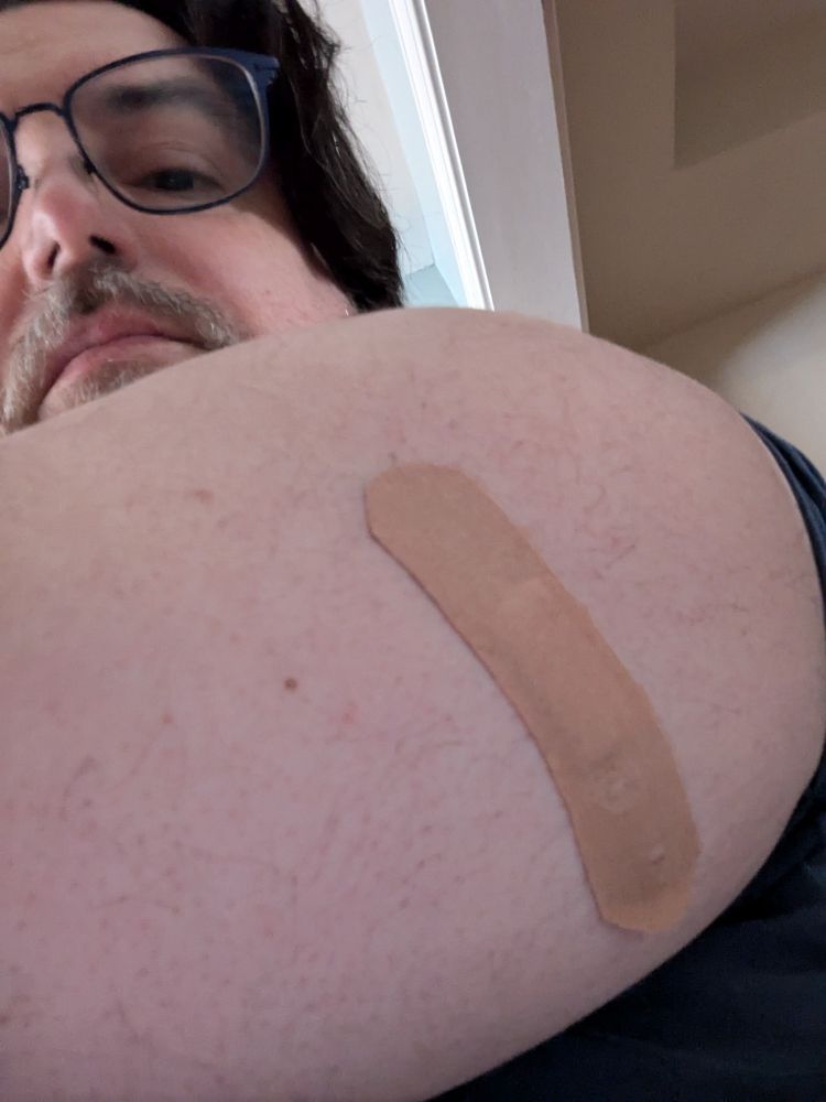 Selfie of a dark-haired, bespectacled, bearded, middle-aged white man. The focus of the image is the back of his upper left arm. An adhesive bandage covers the injection point of an MMR Immunization.