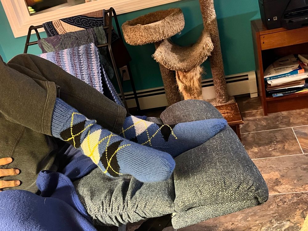 A man’s legs , on a recliner , wearing a pair of argyle socks in blue with black and white diamonds and yellow crossings 