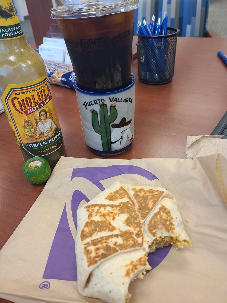 Taco Bell breakfast crunchwrap, green pepper Cholula Hot Sauce, and a cup of cold brew perched on a mug from Puerto Vallarta