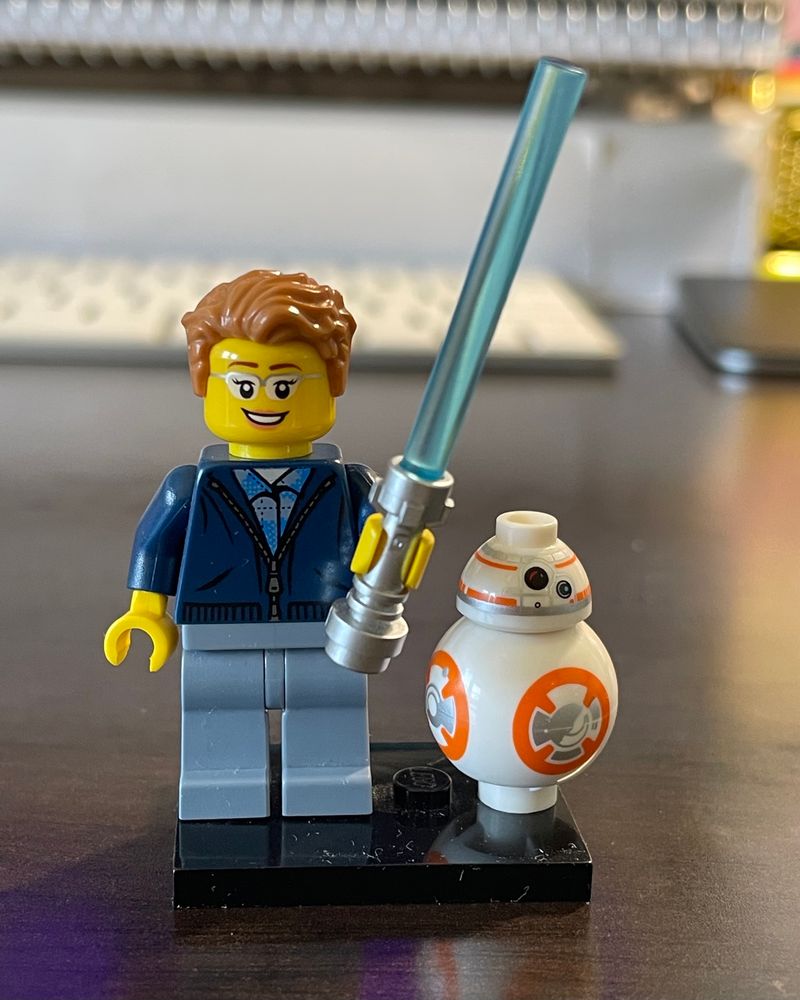 A LEGO minifigure with yellow skin, a smiling female face with glasses, a swoopy blonde pixie cut, a blue zip-up sweater, and gray pants. She’s carrying a metallic blue lightsaber. Beside her stands a small LEGO BB-8.