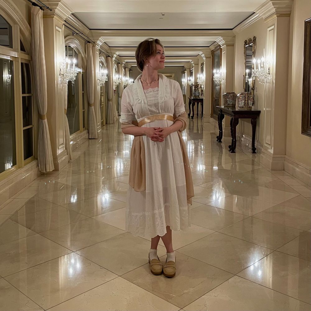 In a vintage hotel hallway, a blonde woman wears a white regency dress with champagne-colored accents.