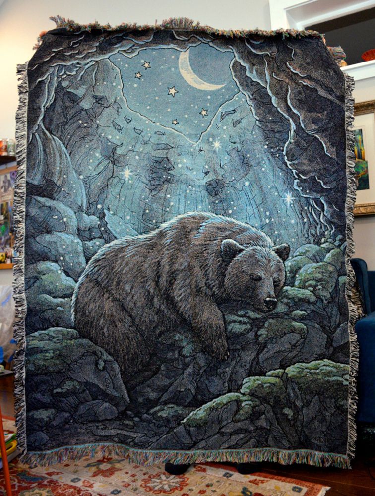 Photograph of a large woven tapestry blanket featuring a brown bear sleeping inside of a mossy cave. There is an opening at the top showing a crescent moon and stars, and sparkles shine down on the bear.