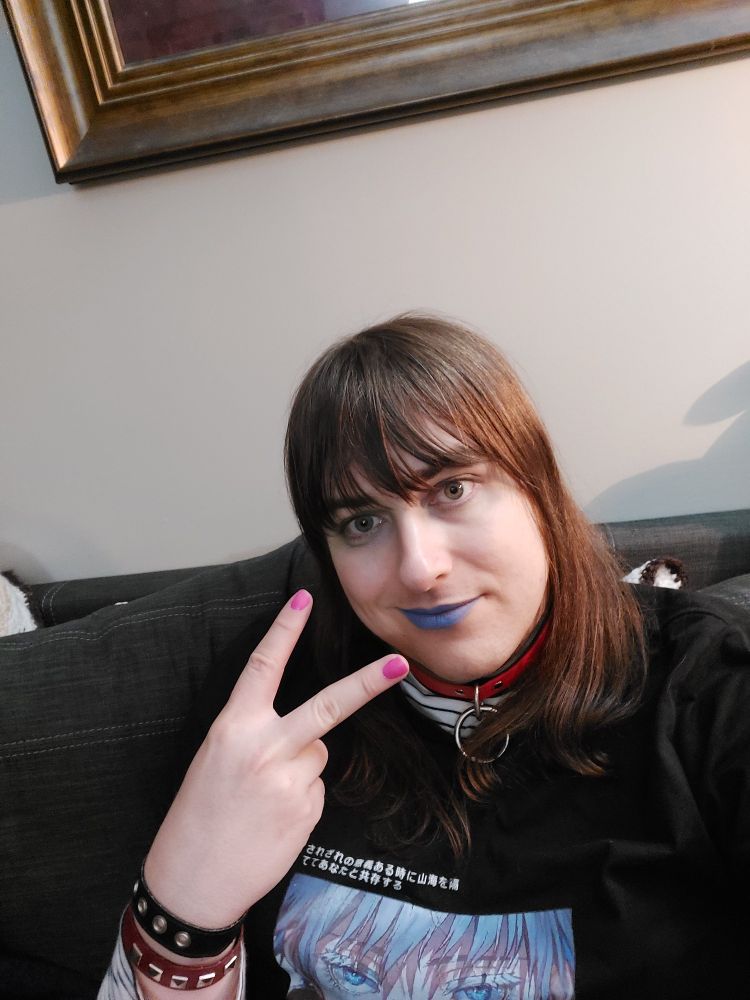 E boi style enby with a collar, multiple wrist bands, an anime t-shirt with a poem in Japanese over a stripped long sleeve shirt, pink nails, and blue lipstick smiling to the camera and showing the peace sign