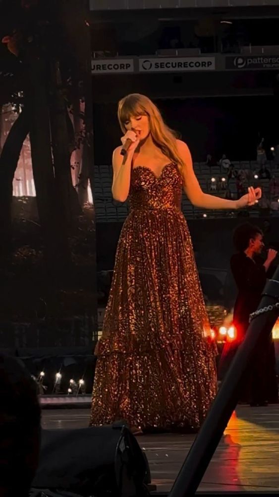 Taylor Swift singing on the Eras tour wearing the bronze evermore dress
