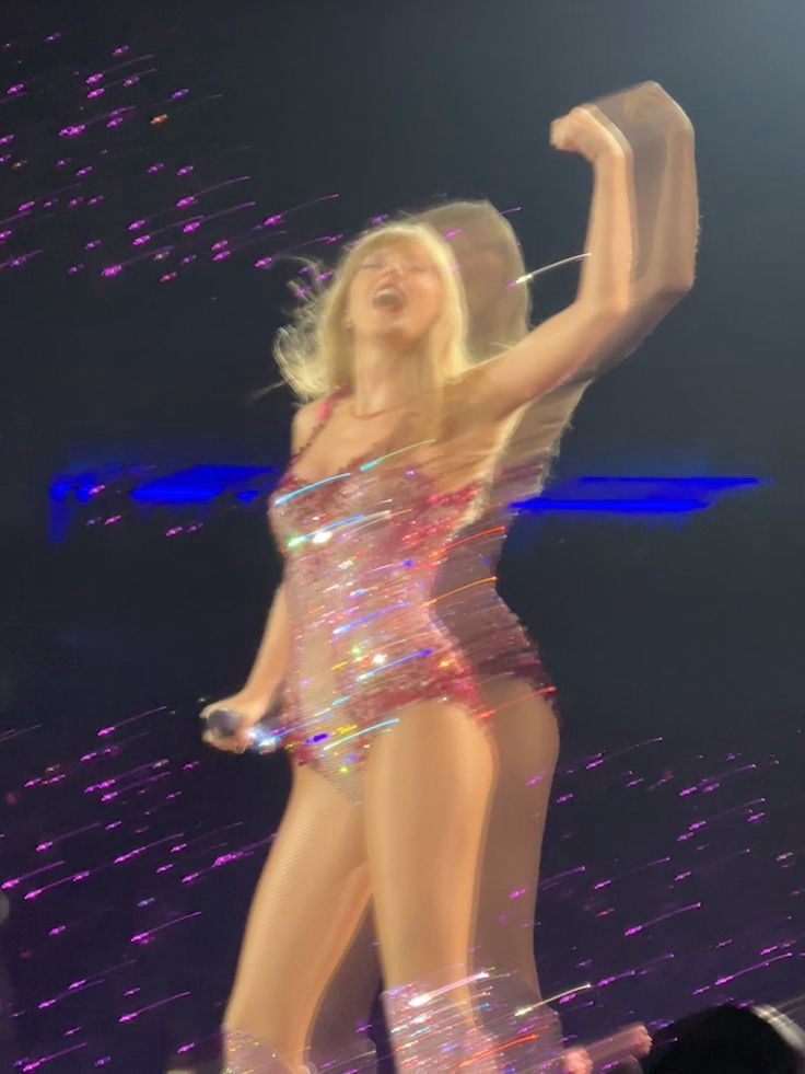 A blurry photo of Taylor Swift on the Eras Tour wearing the pink lover bodysuit with her arm in the air flexing 