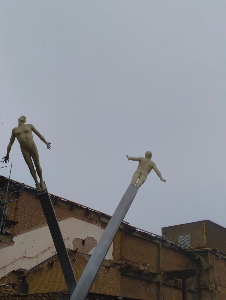 Two stylised figures on posts - one dark, one light.
In the background just below the figures is a partially demolished building