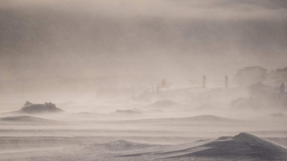 Blowing snow drifting across a frozen cove with houses not clearly visible. Sunlight is dull due to the harsh winds giving a fog like appearance.