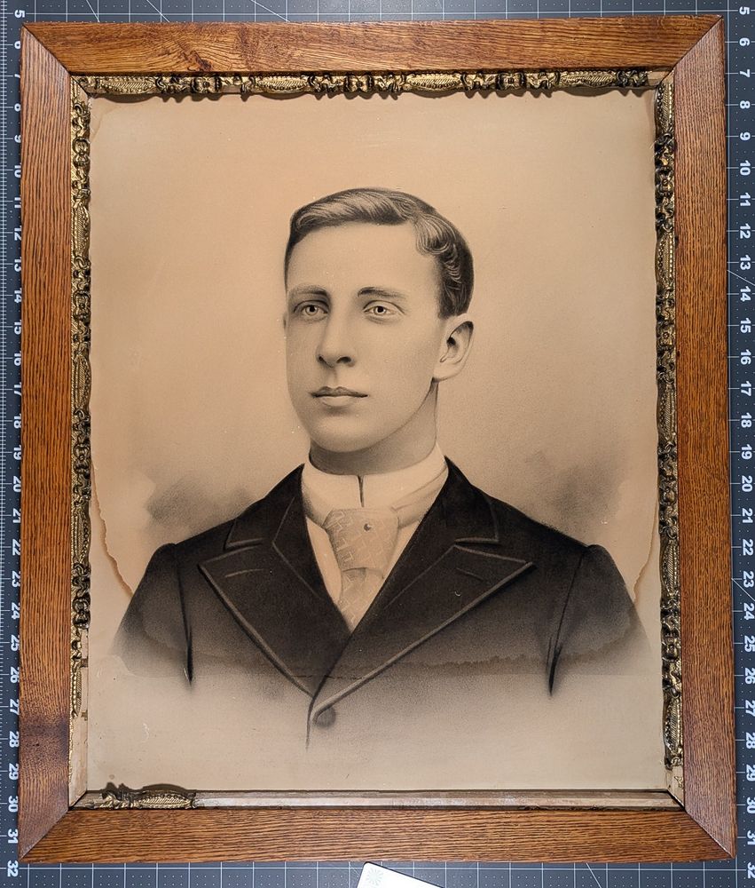 Antique crayon portrait of a young man with a clean shaven face in a decorative frame with some of the decoration pieces missing due to water damage.