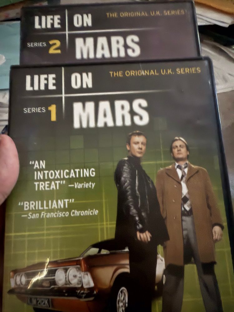 2 DVD covers for the British tv series “Life on Mars: Series 1 & 2.” The photo on the DVD cover is of 2 men standing in front of a 1970s brown car.
