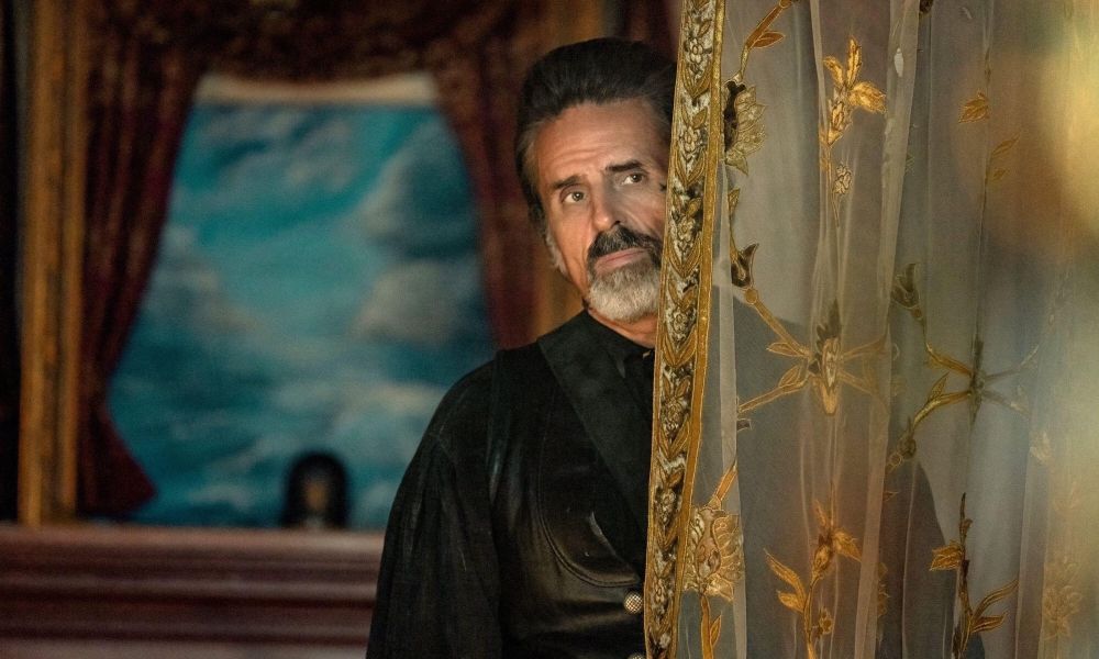 The character Izzy Hands from Our Flag Means Death looking out from behind a thin curtain with gold embroidery. 