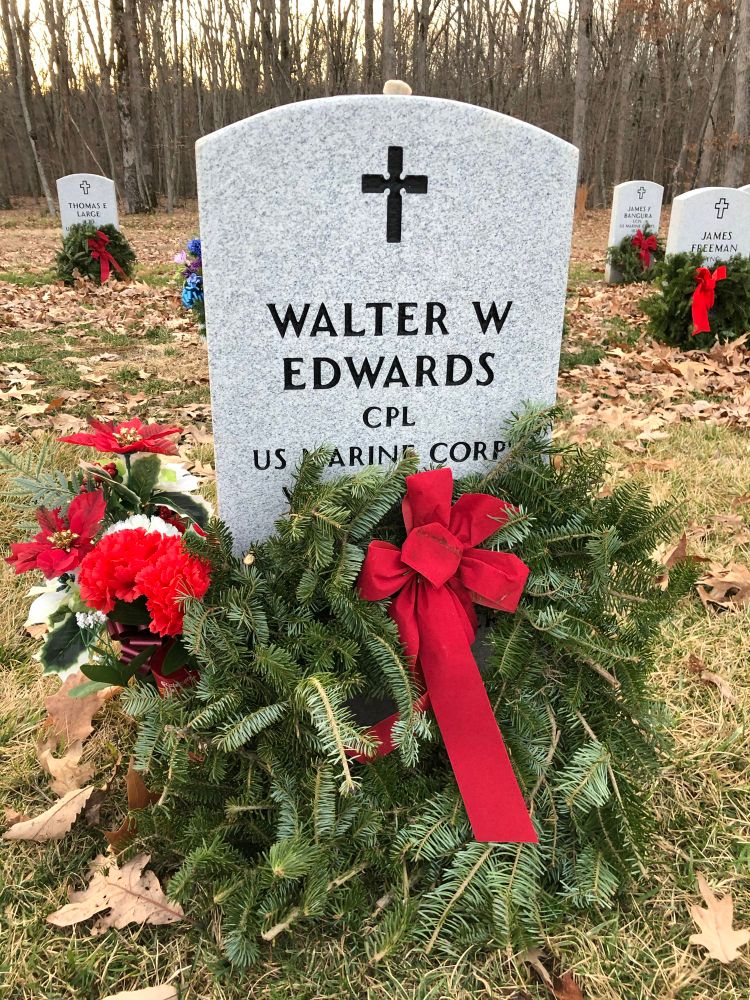 A military headstone at Quantico National Cemetery. A green Xmas wreath with red ribbons sits on the ground in front of the headstone. The headstone says: Walter W Edwards USMC