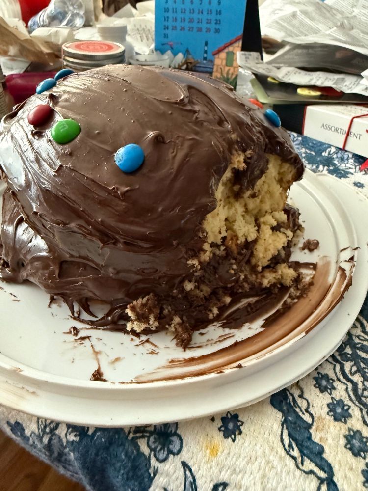 A homemade cake with chocolate frosting. A bit of the right side of the cake has collapsed. 