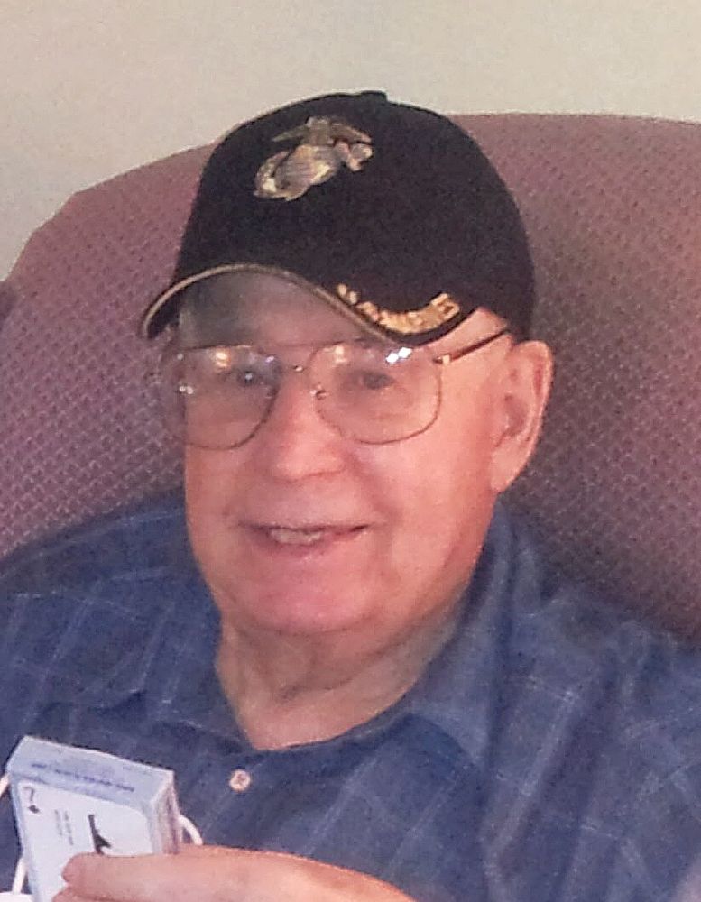 A color photo of a smiling older man who is sitting in a red chair. He has a black baseball style hat on which says “Marines’ along the brim.