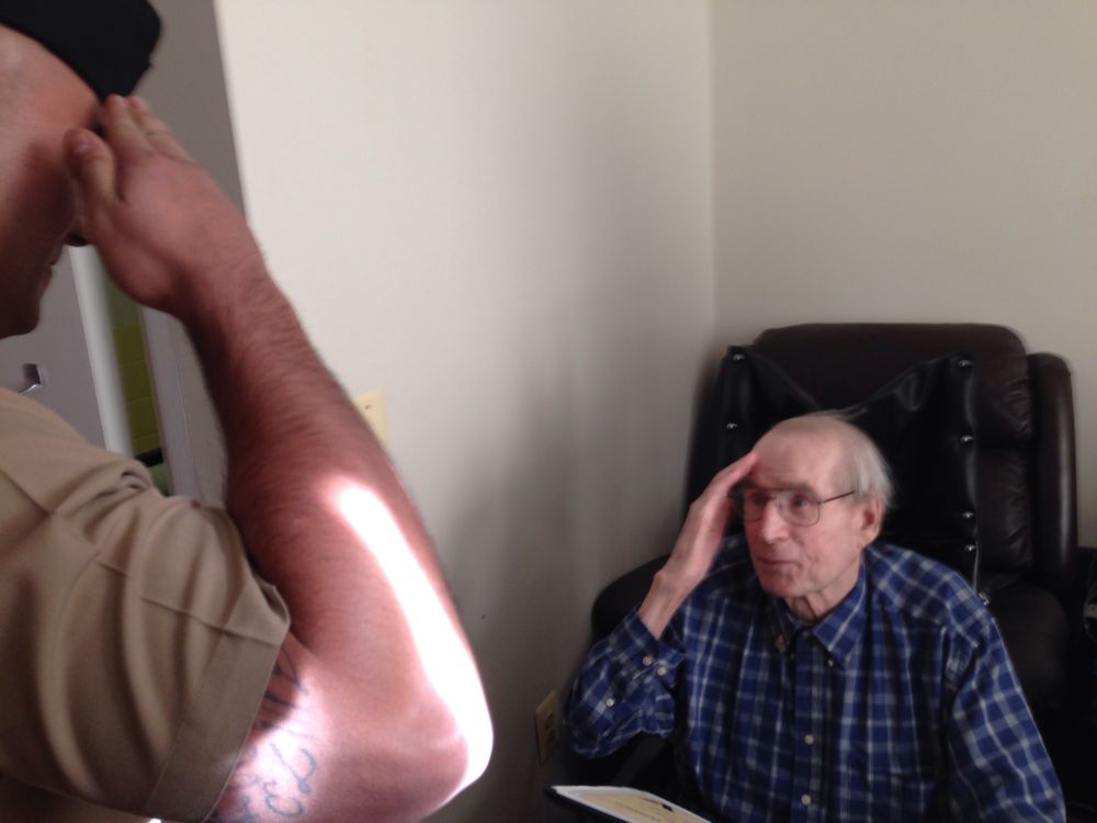 An older man sits in a chair. He is saluting a standing young man wearing a Marine Corps uniform.