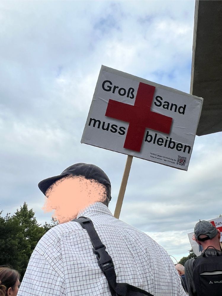 Ein Mann von hinten, der ein Schild mit der Aufschrift „Groẞ-Sand muss bleiben“ und einem roten Kreuz hochhält. Im Hintergrund erahnt man einen Demonstrationszug.