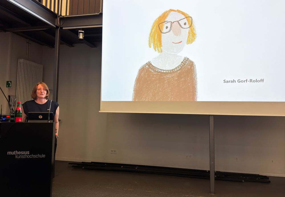 Foto von mir - einer Frau mit roten Haaren und Brille - hinter einem Rednerpult. Neben mir eine große Leinwand, auf der gerade ein Slide mit einem gezeichnetes Selbstporträt von mir zu sehen ist, mit ebensolchen roten Haaren und Brille.