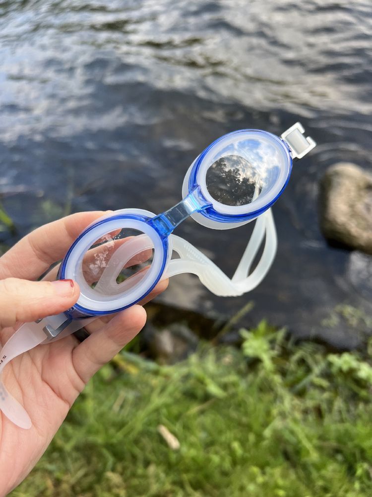 Foto einer Hand, die eine blaue Schwimmbrille hochhält. Im Hintergrund grasbewachsenes Ufer und  See
