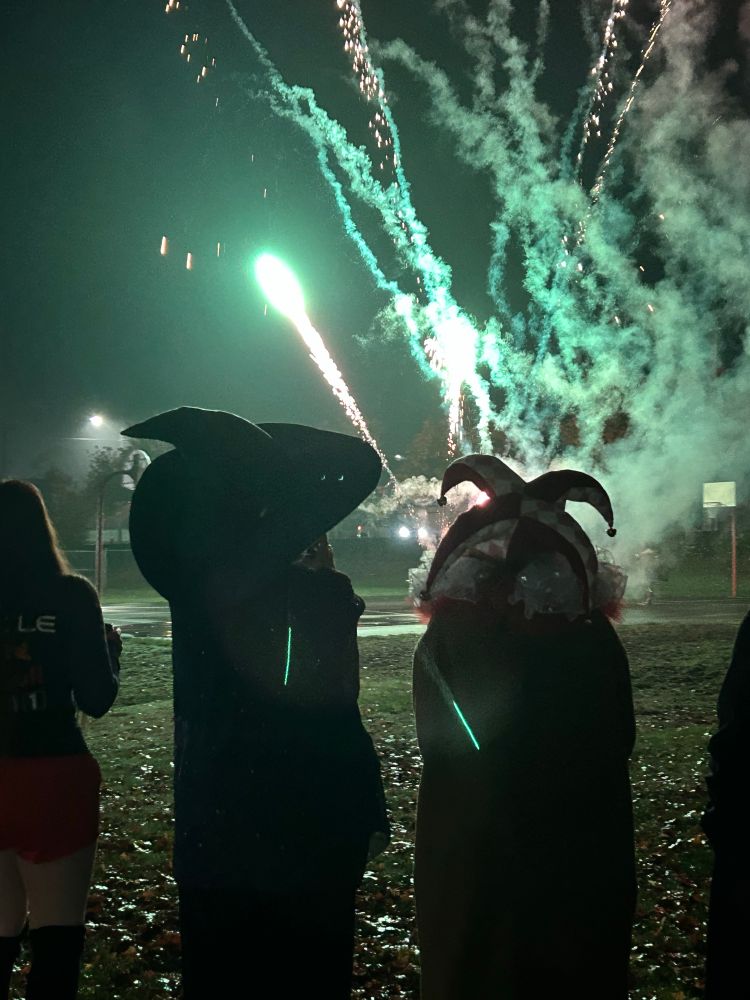 A picture of the back of two figures the left wears a wizard hat and robes and the right wears a four pointed jesters hat and a ruffled collar they are dark silhouettes in the night. In front of them are fireworks being lit that let off an eerie light teal glow 