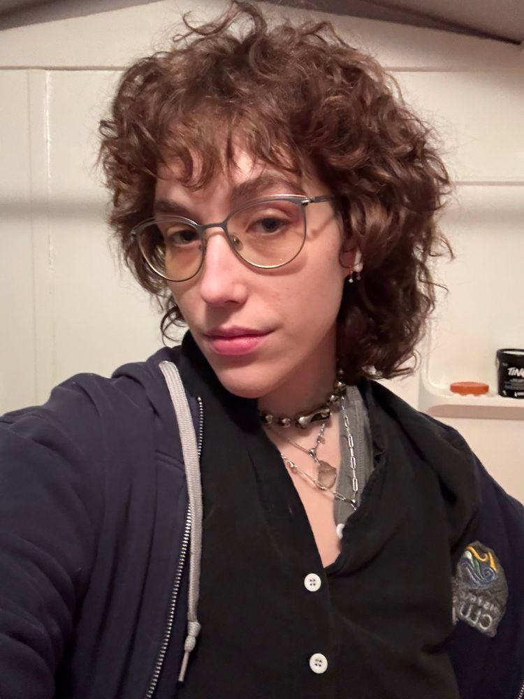 Selfie of Shelby a white nonbinary person with short curly auburn hair, glasses and wearing multiple necklaces. They are looking toward the camera with their face turned. They aren’t pretending to like how they look. 