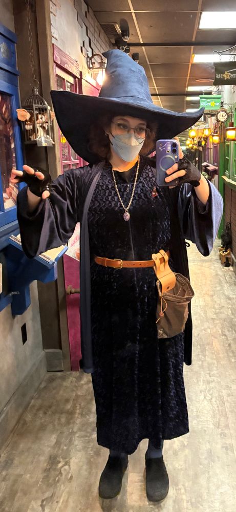 Shelby a white person infront of a floor length mirror wearing blue velvet robes and a blue wizard hat and a blue mask to protect themselves and their community from COVID. They have a spell pouch and a powerful amulet around their neck 