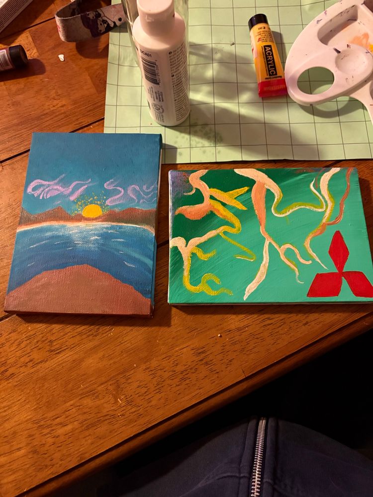 Picture of two small paintings in a wooden table. Left painting is vertical while the right is horizontal. The left depicts a seaside cliff looking out over the sea to a mountain range with a setting/rising sun depending on the perspective. Often used in a Wands card. 

The right is a green gradient background with a Mitsubishi symbol that I saw in a dream and then these like fleshy toned roots/squiggles. Maybe it means something about humans and brands and how we attribute so much of our lives to them when it means nothing. Who knows. 