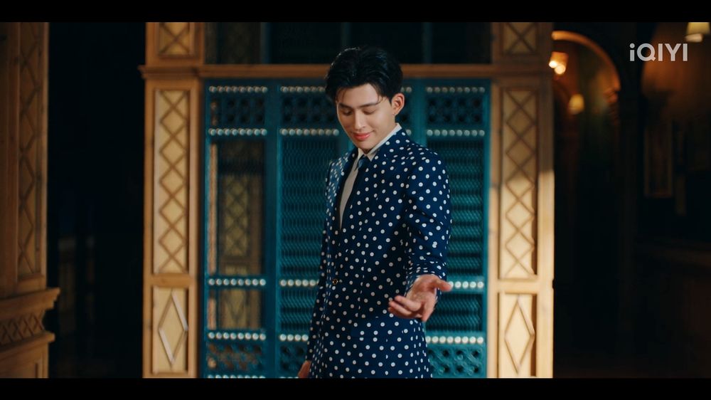 a screenshot from episode 2 of the Thai BL drama "Me and Thee"

Thee (played by Pond) is wearing a blue suit with white polka dots, a blue tight, and white shirt. he is standing with his body turned to the left, but his head forward, eyes to the ground. he is holding out his left arm in invitation. the set behind him shows elegant molding and decor, in natural wood and a deep teal/green tone.