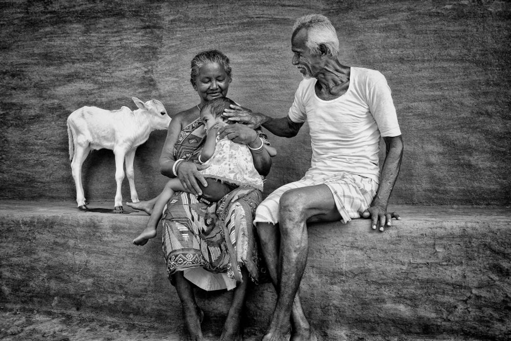 Image by Tanusree Mitra. A black and white image of an elderly couple in Purulia, West Bengal, India, as they sit on a ledge with their young granddaughter. The grandmother cradles the child, who rests tiredly on her lap, while the grandfather lovingly strokes the child’s hair. A white calf stands beside them, adding to the serene and intimate rural scene.