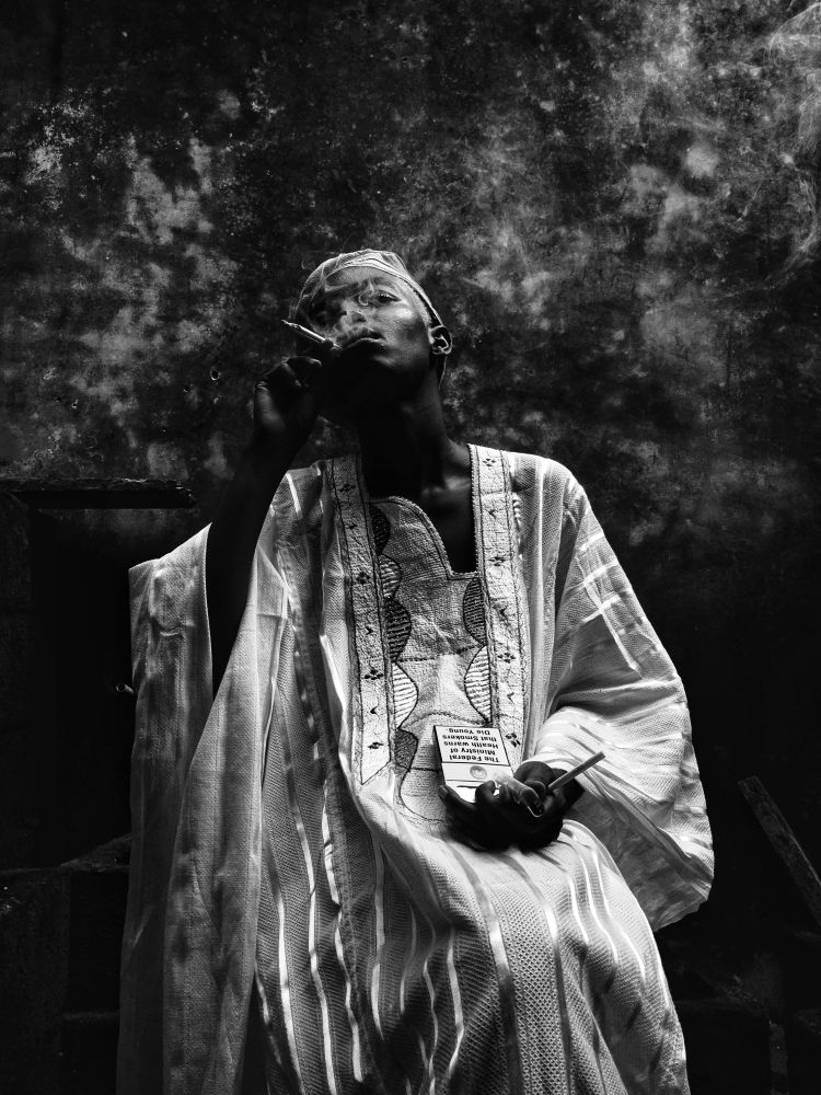 Black-and-white portrait of a woman in traditional agbada and cap, smoking a cigarette and holding a drink; dramatic lighting enhances the contrast and mood, evoking the visual style of vintage tobacco and alcohol ads.