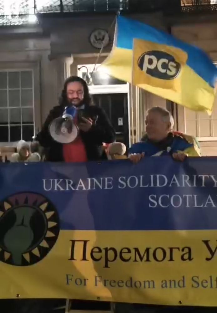 Vijay standing with megaphone behind USC Scotland banner outside US Consulate, Edinburgh. PCS Union and Ukraine flags wave in background. 